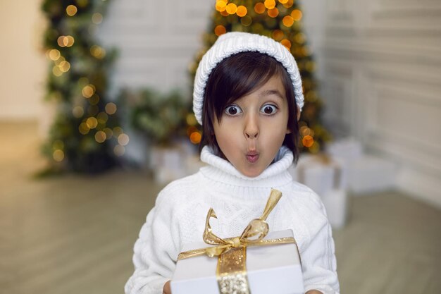 Garçon dans un pull en tricot blanc et un chapeau se tient avec une boîte-cadeau à l'arbre de Noël à la maison le jour de Noël