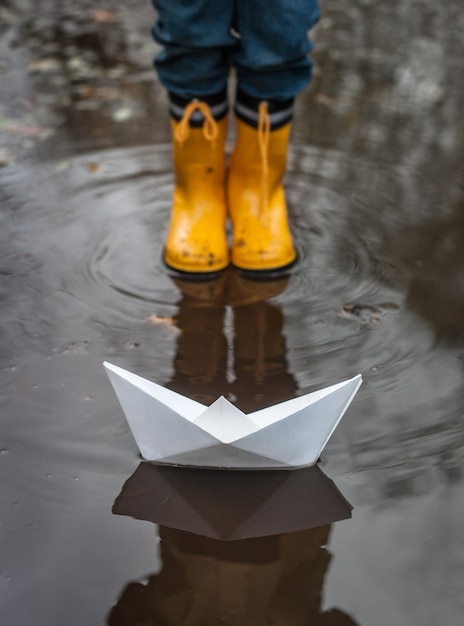 Un garçon debout devant un bateau en papier dans une flaque d'eau.