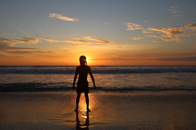 Un garçon debout sur la plage contre le ciel au coucher du soleil