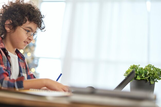 Garçon de l'école latine enthousiaste portant des lunettes à faire ses devoirs à l'aide d'une tablette numérique assis à la