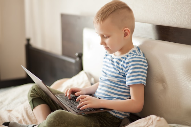 Garçon enfant assis sur le lit et à l'aide d'un ordinateur portable numérique