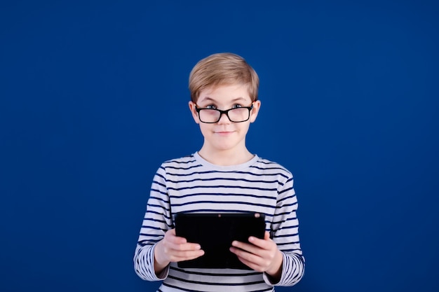 Garçon enfant blond avec de grandes lunettes jouant avec tablette sur mur bleu. Concept de l'éducation.