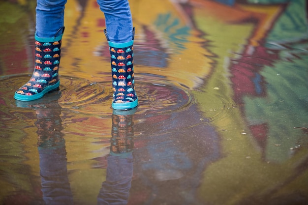 Garçon enfant dans des bottes en caoutchouc drôles debout dans la flaque d'eau dans la rue après la pluie