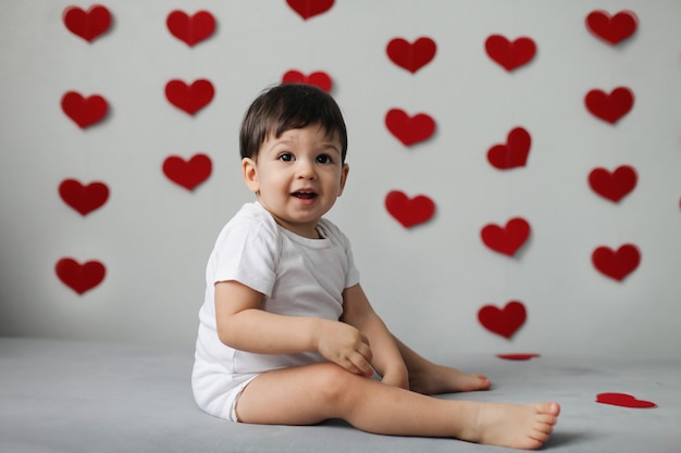 Garçon enfant heureux dans un body blanc avec noeud papillon se trouve sur un fond d'un mur gris avec des coeurs le jour de la Saint-Valentin.