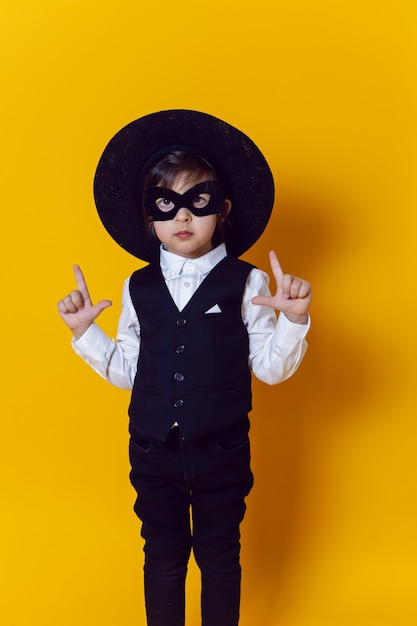 Garçon enfant super héros dans un costume et un masque noir et un pistolet chapeau sur un mur jaune pieds nus
