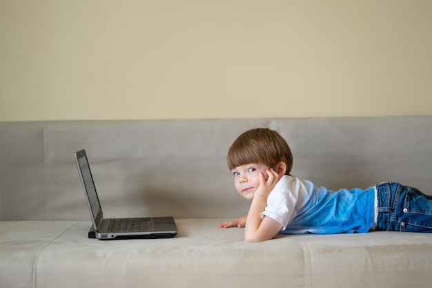 Le garçon est allongé sur un canapé lumineux et regarde un ordinateur portable. L'enfant communique avec ses amis par