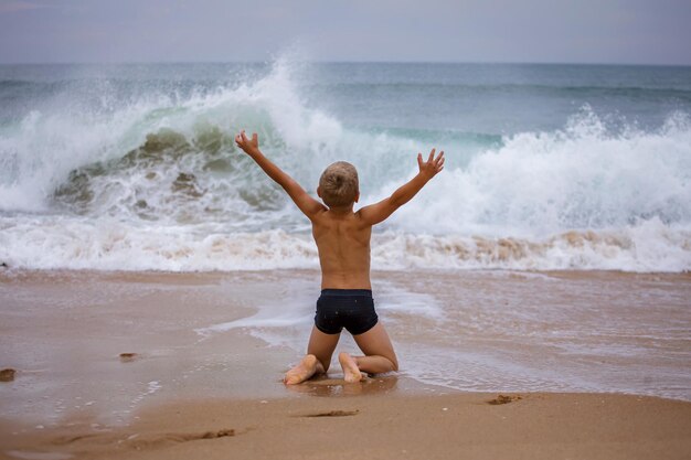 Photo garçon est assis sur le rivage de l'océan avec ses bras ouverts vers le vent et les vagues tempête en été courage