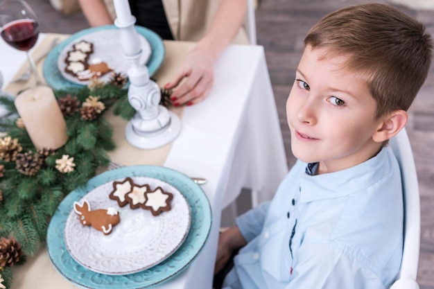 Le garçon est assis à la table de Noël et regarde la caméra