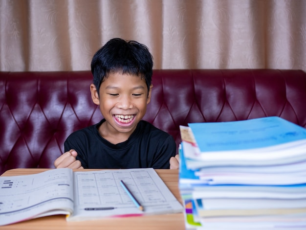 Le garçon était très heureux de finir ses devoirs. Il était assis sur une table en bois et avait une pile de livres à côté. L'arrière-plan est un canapé rouge et des rideaux crème.