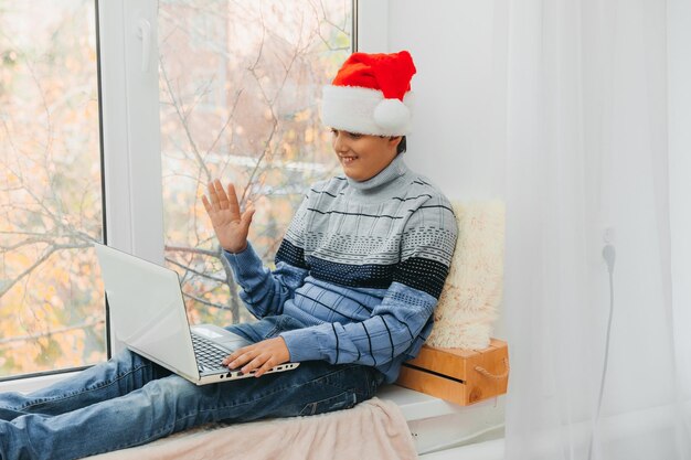 Un garçon européen portant un chapeau de Père Noël est assis sur le rebord de la fenêtre près de la fenêtre avec un ordinateur portable et communique par liaison vidéo.