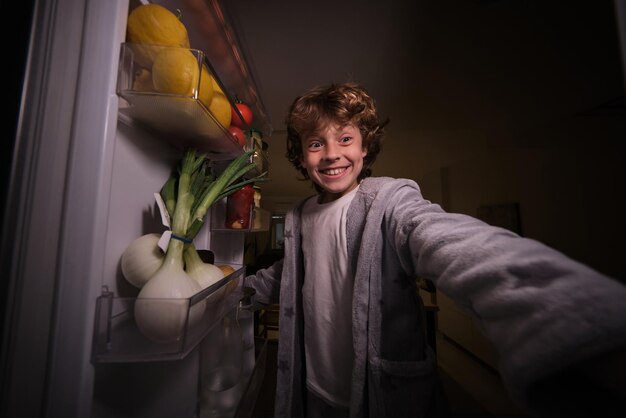 Garçon excité en vêtements de nuit debout près de la porte ouverte du réfrigérateur avec divers produits à manger tout en choisissant de la nourriture la nuit