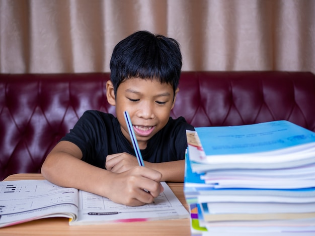 Garçon faisant ses devoirs et lisant sur une table en bois avec une pile de livres à côté L'arrière-plan est un canapé rouge et des rideaux crème.