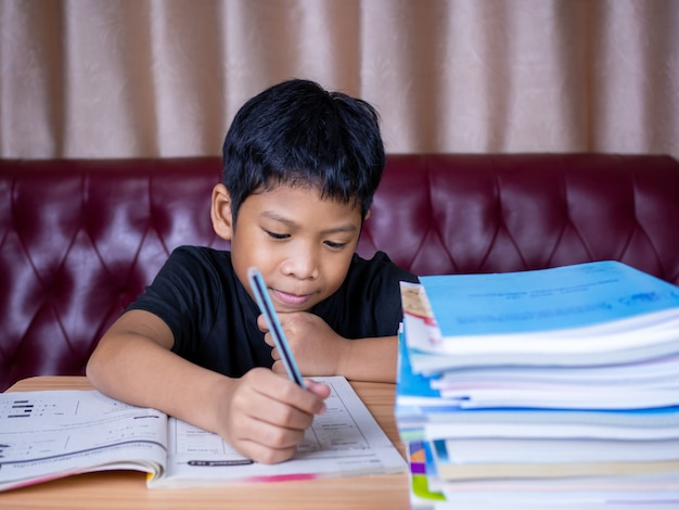 Garçon faisant ses devoirs et lisant sur une table en bois avec une pile de livres à côté L'arrière-plan est un canapé rouge et des rideaux crème.