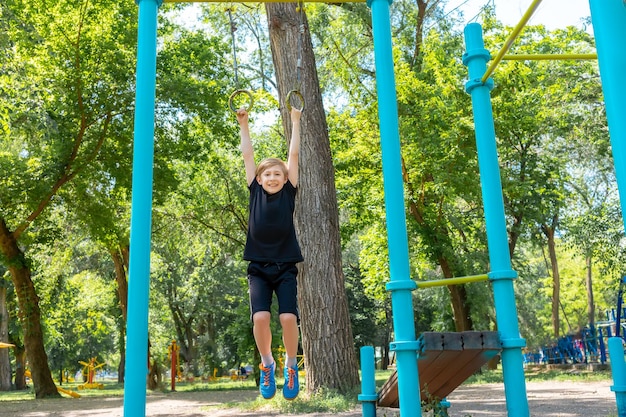 Le garçon fait du sport dans le parc, il s'accroche aux anneaux de gymnastique et essaie de se relever