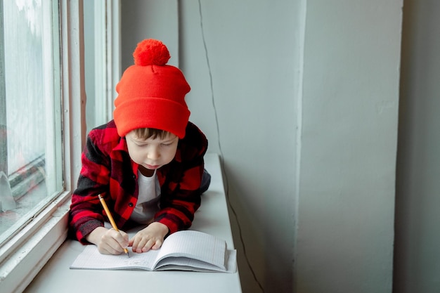 Le garçon fait ses devoirs Le garçon est allongé sur le rebord de la fenêtre et écrit dans un cahier Écolier fait ses devoirs près de la fenêtre Enseignement à domicile