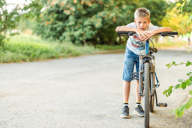 Un garçon fatigué dans un t-shirt et un short en jean s'appuie sur son vélo après un long trajet à vélo dans le parc