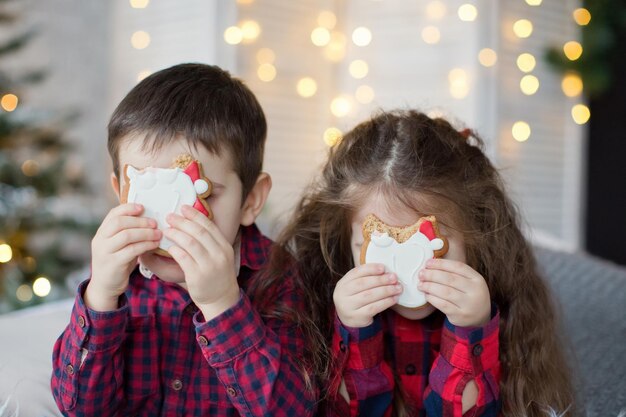 Un garçon et une fille en bas âge en chemises rouges mangent des biscuits près de l'arbre de Noël