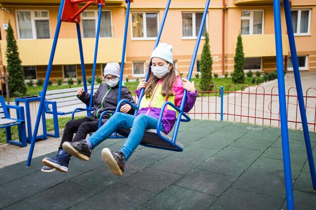 Le garçon et la fille de l'école élémentaire portent un masque médical et se balancent sur une balançoire Les enfants sérieux se reposent dans la cour de récréation à l'automne