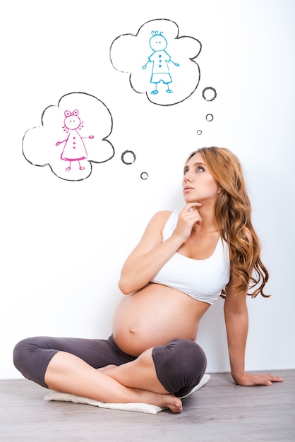 Garçon ou fille? Femme enceinte réfléchie levant et tenant la main sur le menton tout en étant assise sur fond blanc