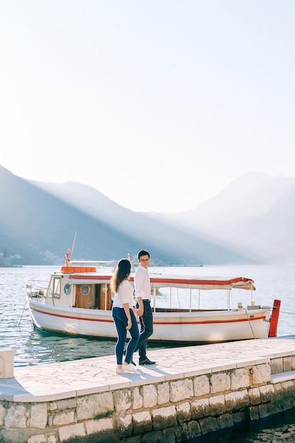 Un garçon et une fille se tiennent la main le long de la jetée devant un bateau amarré.