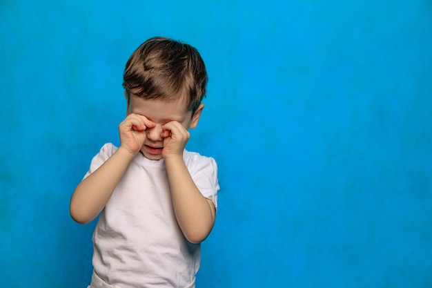 Un garçon sur fond bleu se frotte les yeux. Santé des yeux. Maladies des yeux. Un bébé qui pleure. Fatigue des enfants. Psychologie.