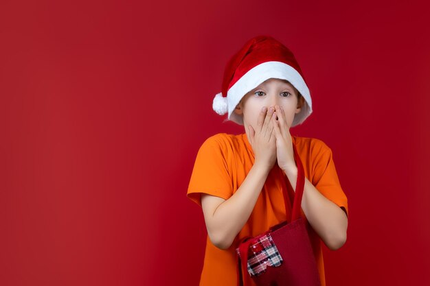 Un garçon sur fond rouge dans un chapeau de père Noël tient un sac-cadeau dans ses mains et est surpris