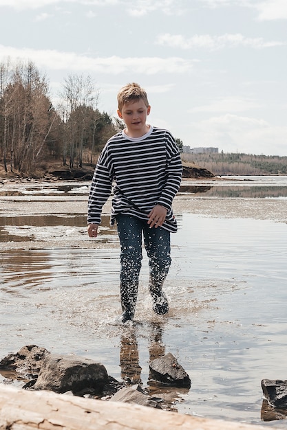 Un garçon gai dans un gilet rayé et des bottes en caoutchouc saute dans l'eau sur la rive du fleuve.