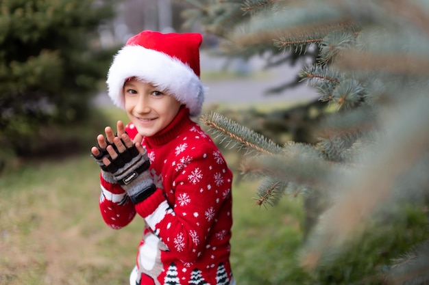 Un garçon gai dans un pull de Noël rouge avec un renne et un bonnet de Noel se frotte les mains et rit malicieusement