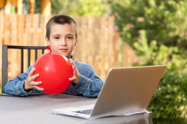 Un garçon gonfle un ballon rouge par une chaude journée d'été dans le jardin. Préparation d'anniversaire