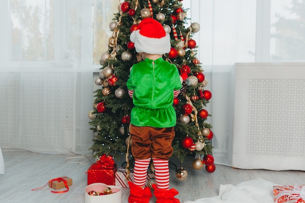 Un garçon habillé en gnome et portant un chapeau du Nouvel An décore un arbre de Noël à la maison. Noël.