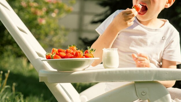 Un garçon de huit ans mange des fraises avec du yaourt en été dans la cour