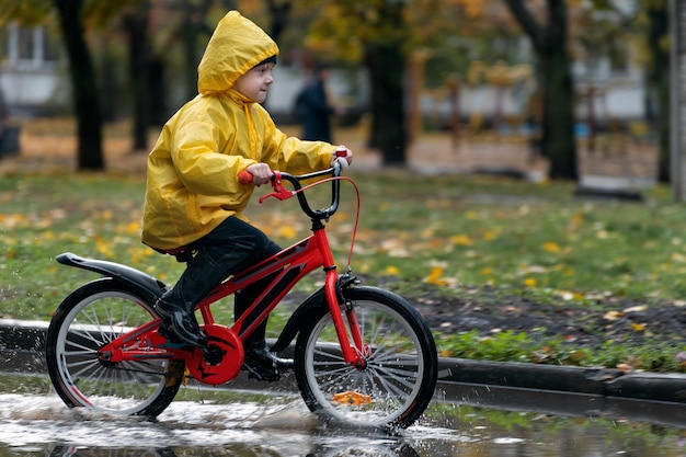 Garçon en imperméable est seul à vélo à travers les flaques d'eau. Un enfant heureux fait du vélo sous la pluie.