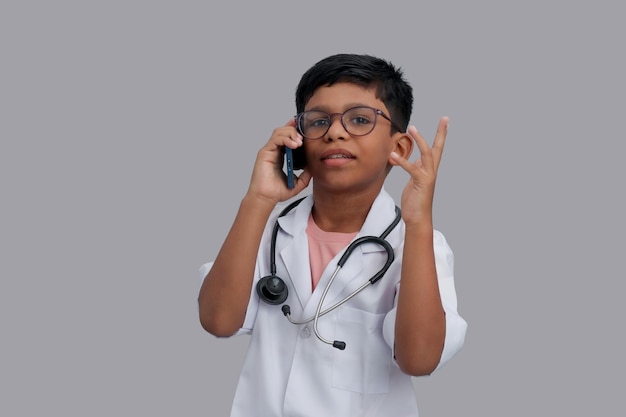 Un garçon indien âgé de 7 à 8 ans portant un tablier de médecin avec un stéthoscope qui parle sur un smartphone