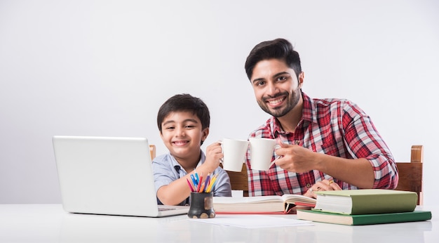 Garçon indien mignon avec père ou tuteur masculin faisant ses devoirs à la maison à l'aide d'un ordinateur portable et de livres - concept de scolarisation en ligne