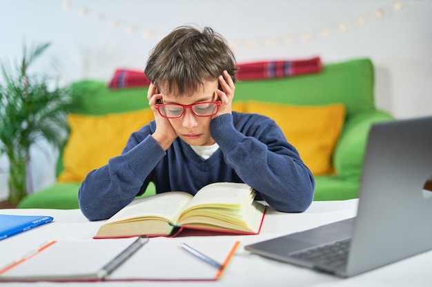 Un garçon intelligent concentré avec les mains sur la tête lit un livre à l'école avec des cours en ligne à la maison en quarantaine,