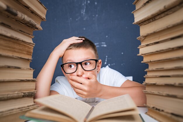 Garçon intelligent dans des verres devenir fou de lire un livre alors qu'il était assis entre deux piles de livres