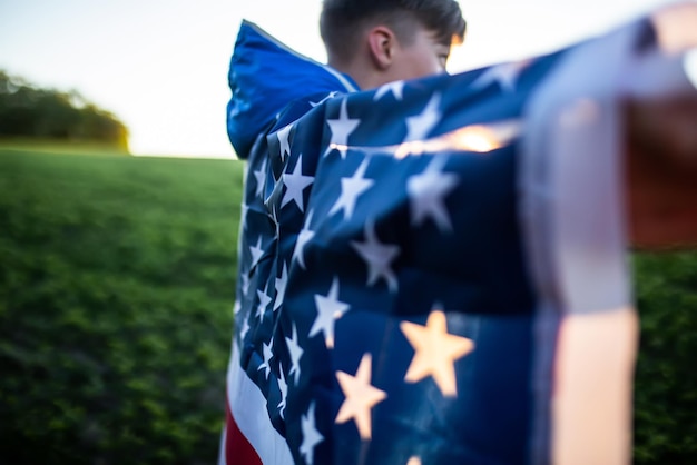 Un garçon a jeté le drapeau de l'Amérique sur ses épaules. Le drapeau des États-Unis au coucher du soleil à l'extérieur le jour de l'indépendance.