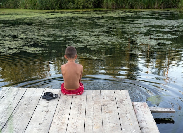 Garçon sur la jetée regardant l'eau garçon assis sur une jetée en bois vue de l'arrière sur la rivière calme et silence au coucher du soleil