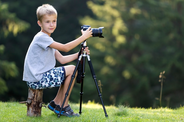 Garçon jeune enfant blond assis sur une souche d'arbre sur une clairière prenant une photo avec caméra trépied.