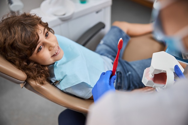 Garçon joyeux assis dans un fauteuil dentaire en regardant son dentiste démontrant la bonne façon de se brosser les dents avec un modèle de mâchoires