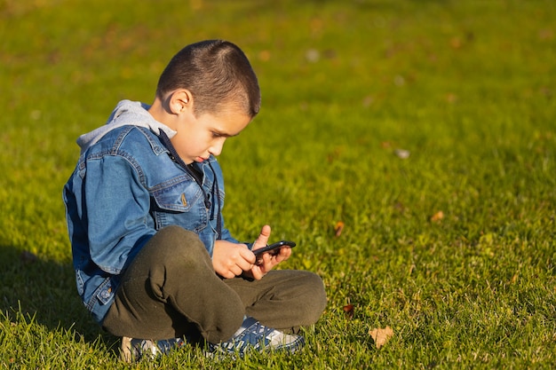 Un garçon joyeux dans une veste en jean est assis sur une pelouse verte et joue avec un smartphone dans un parc de la ville