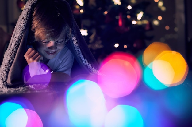 Garçon joyeux lisant un livre intéressant avec une lampe de poche lumineuse tout en étant allongé sur le lit dans une pièce sombre avec un bokeh coloré à la maison