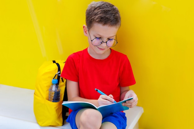 Un garçon joyeux avec des lunettes est assis sur un banc près de l'école et écrit dans un cahier