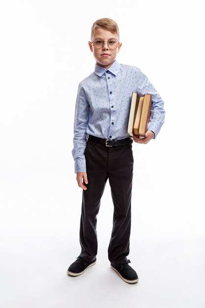 Garçon à lunettes avec des livres dans ses mains. Enfant de 9 à 10 ans en chemise et pantalon bleus. Éducation et étude. Pleine hauteur. Fond blanc. Verticale.