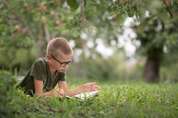 Un garçon avec des lunettes se trouve sur l'herbe dans le parc et lit un livre