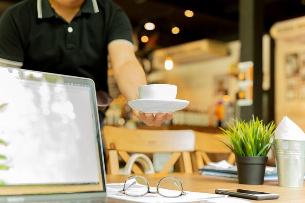 Garçon masculin servant du café avec un ordinateur portable et des verres sur la table de travail au café.