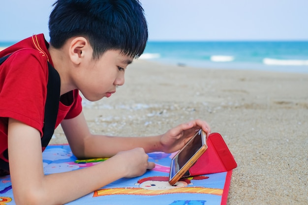 Garçon mignon asiatique à l'aide de tablette pour jouer à un jeu sur la plage