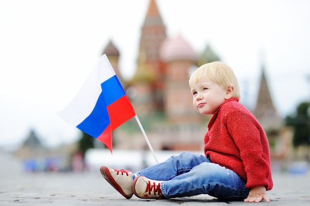 Garçon mignon bambin assis et jouant avec le drapeau russe avec la place rouge et la descente de Vasilevsky