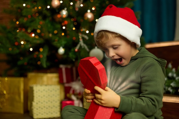 Le garçon mignon dans un chapeau de Noël ouvre un cadeau magique se reposant à un arbre de Noël décoré