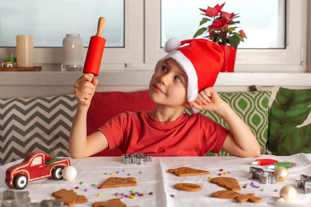 Un garçon mignon dans un chapeau de père Noël fait du pain d'épice à la table et tient un rouleau à pâtisserie rouge dans ses mains